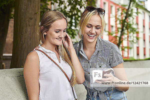 Two happy young women sharing cell phone and earphones outdoors
