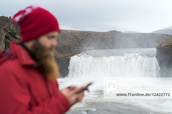 Iceland  North of Iceland  young man using smartphone  waterfall in the background