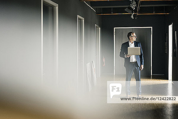 Mature businessman standing on office floor holding laptop