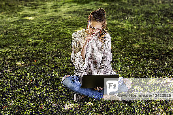 Pensive woman sitting on a meadow using laptop