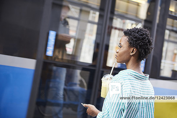 Woman with smoothie and smartphone waiting at bus stop