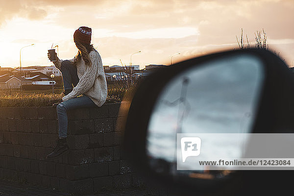 Iceland  young woman with coffee to go at sunset  wing mirror
