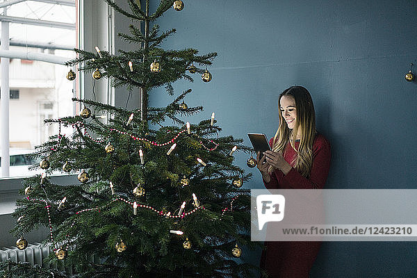 Smiling woman standing besides decorated Christmas tree using tablet