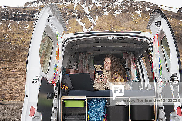 Portrait of young woman lying in van using smartphone and laptop