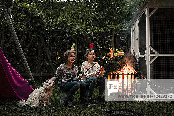 Two girls wearing feather headdress  grilling sausage over camp fire  dog sitting on meadow