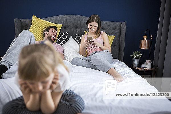Neglected little girl sitting on bed with her parents  using smartphones