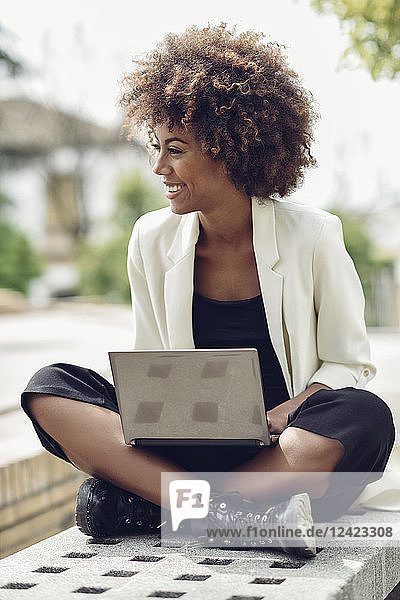 Fashionable young woman with curly hair sitting on bench with laptop laughing