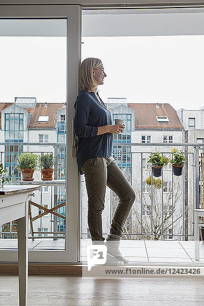 Smiling woman holding cup of coffee looking out of balcony door