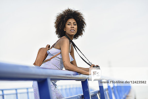 Portrait of fashionable young woman with backpack and camera standing on a bridge