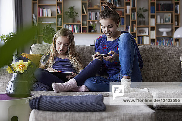 Two sisters sitting on sofa  playing and catting with digital devices