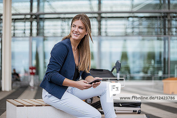 Smiling young businesswoman sitting outdoors with cell phone and suitcase looking around