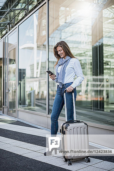 Smiling young businesswoman with suitcase looking at smartphone