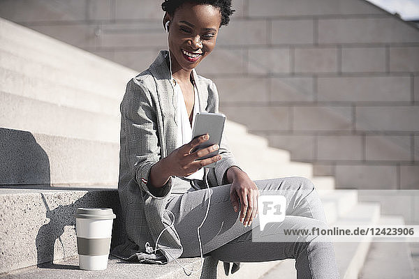 Portrait of smiling businesswoman sitting on stairs using earphones and smartphone