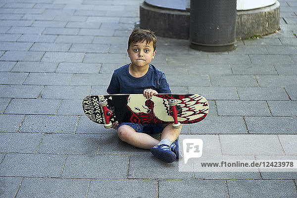Portrait of astonished little boy sitting on pavement with skateboard
