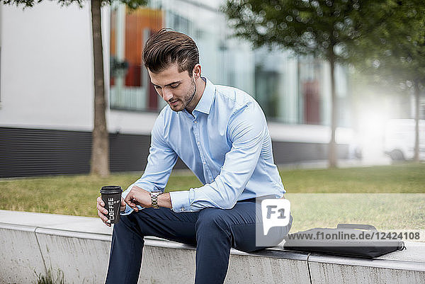 Young businessman sitting outdoors with takeaway coffee checking the time