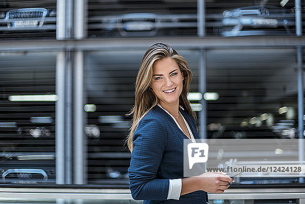 Portrait of smiling young businesswoman with cell phone in front of blurred parking garage