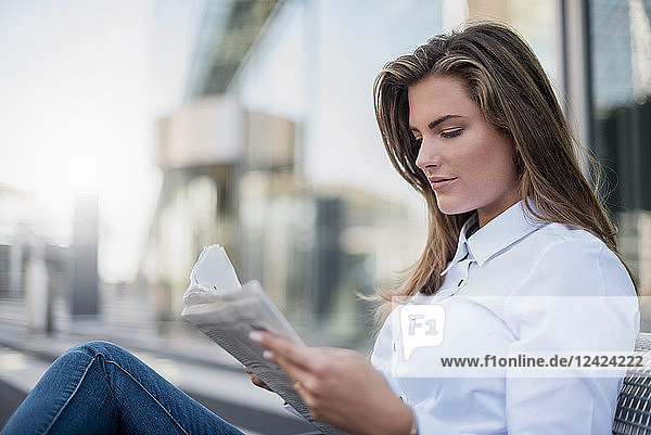 Portrait of young businesswoman sitting on bench reading newspaper