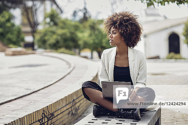 Fashionable young woman with curly hair sitting on bench with laptop