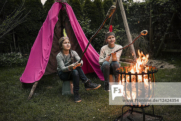 Two girls wearing feather headdress  with stock bread at camp fire