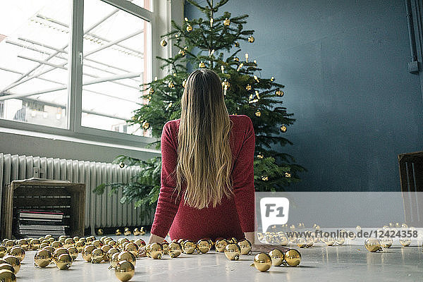 Back view of woman sitting on the floor with many golden Christmas baubles looking at Christmas tree