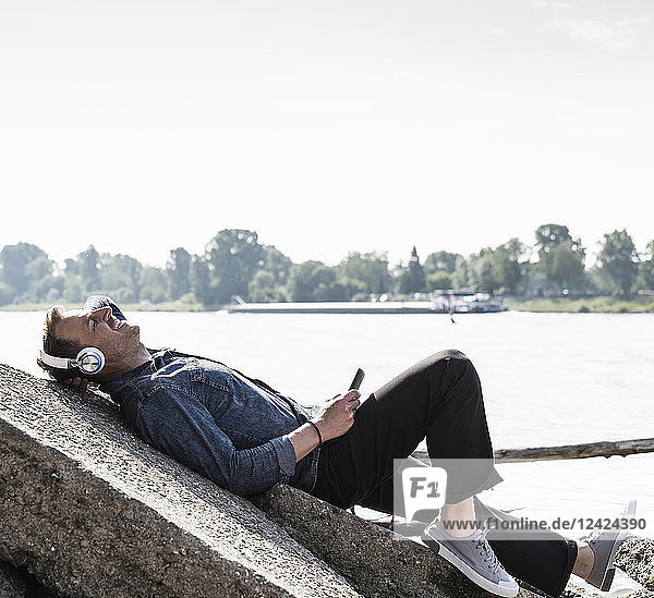 Mature man with headphones and smartphone at Rhine riverbank