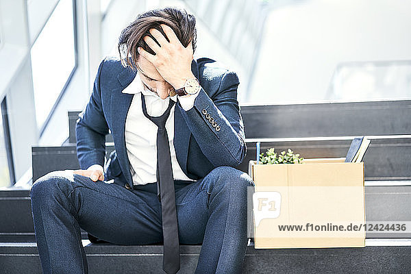 Depressed businessman sitting on stairs with belongings in cardboard box beside him