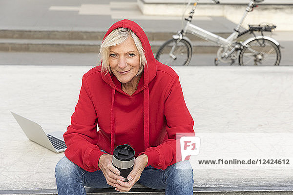 Senior woman wearing red hoodie sitting outdoors with laptop and takeaway coffee