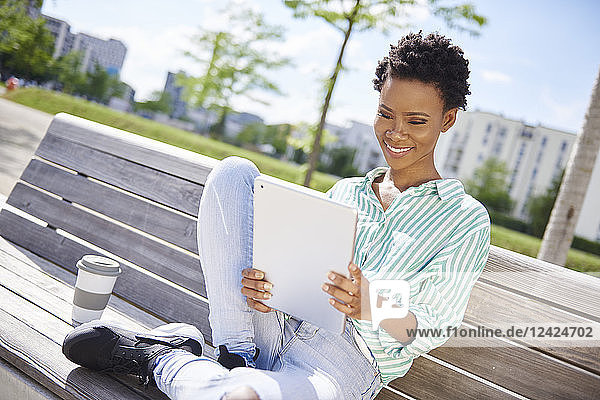 Portrait of smiling young woman with coffee to go sitting on bench looking at tablet