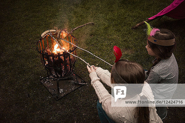 Two girls wearing feather headdress  grilling sausage over camp fire