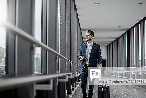 Young businessman in a passageway with cell phone  earbuds and rolling suitcase on the go