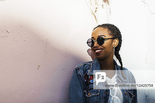 Lächelnde junge Frau mit Sonnenbrille und Jeansjacke vor einer weißen Wand