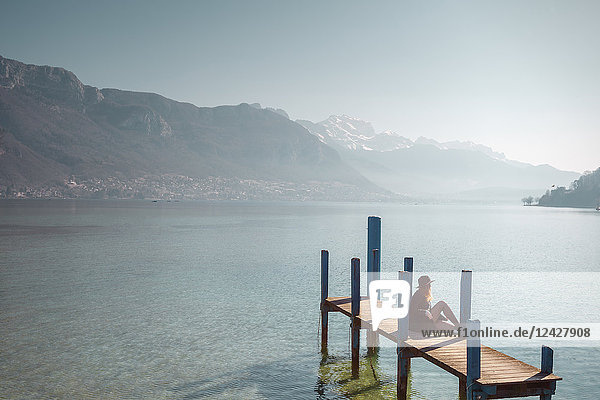 Frau sitzt auf einem Steg am Seeufer unter klarem Himmel mit Bergen im Hintergrund  Annecy  Haute-Savoie