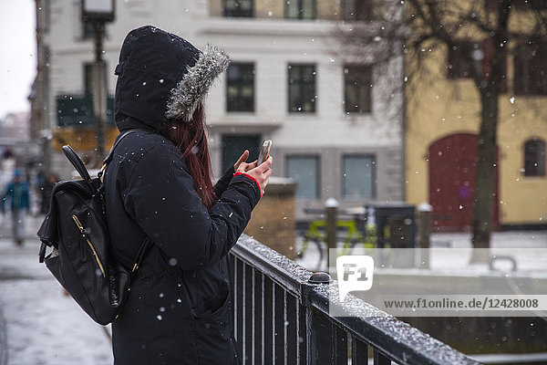 Seitenansicht einer Frau mit Kapuzenjacke und Rucksack  die ein Foto mit ihrem Smartphone auf einer Brücke in Kopenhagen  Dänemark  macht