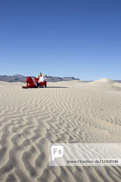 Santa Claus drinking beer while lying on sand in desert under clear sky
