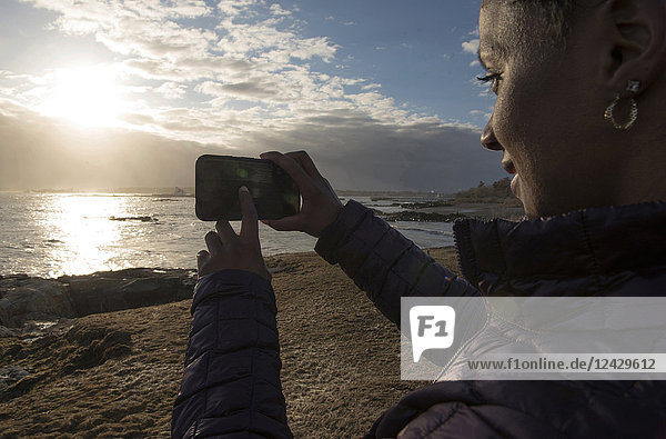 Blick über die Schulter auf eine einzelne afroamerikanische Frau  die ein Foto mit ihrem Smartphone am Meer bei Sonnenuntergang macht