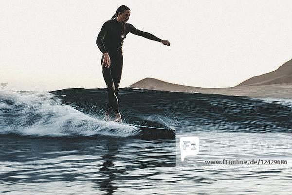 Full length shot of surfer in wetsuit riding wave in sea at sunset