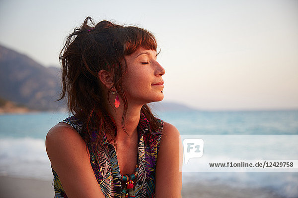 Kopf und Schultern Porträt der schönen jungen Frau mit geschlossenen Augen am Strand bei Sonnenuntergang