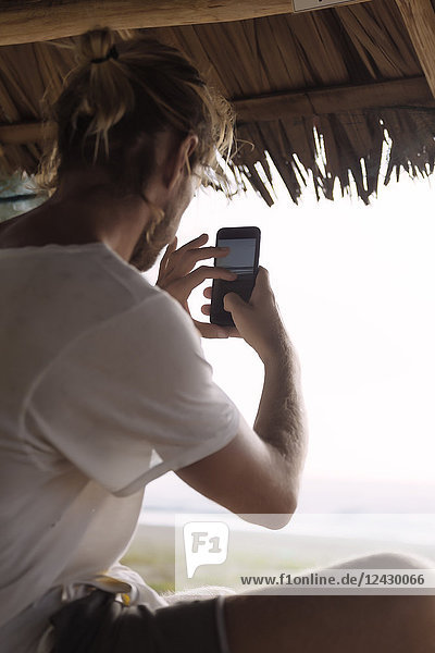 Mann sitzt unter einem Strohzelt am Strand und fotografiert das Meer mit seinem Smartphone  Banda Aceh  Sumatra  Indonesien