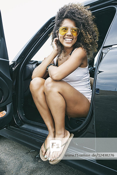 Portrait of smiling young woman with brown curly hair sitting in car  wearing sunglasses  white vest and flip flops.