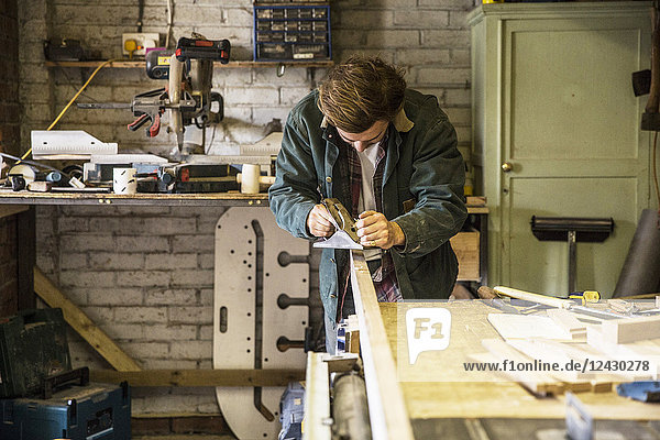 Mann  der in einer Holzwerkstatt stehend an einer Werkbank arbeitet  mit einem Hobel auf einem Brett aus Holz.