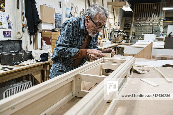 Senior Caucasian carpenter sanding a wooden cabinet part in a large woodworking shop.