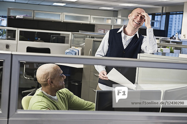 Caucasian male and black male talking and smiling in a cubicle office setting.