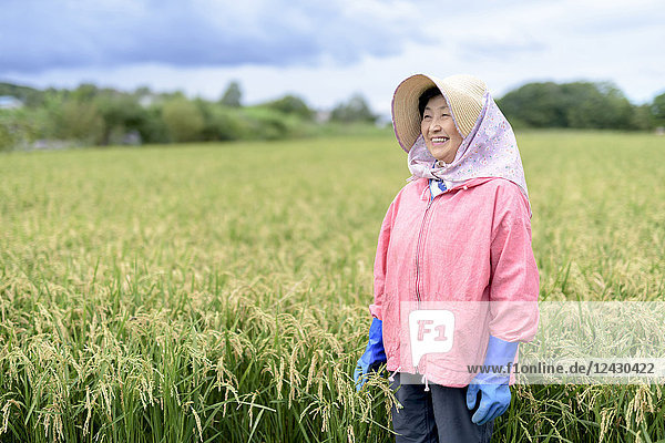 Lächelnde Frau mit Strohhut  rosa Jacke und blauen Gummihandschuhen steht in einem Reisfeld.