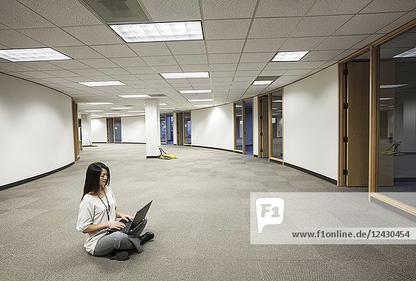 An Asian businesswoman sitting cross legged and working on a laptop computer in the middle of an empty office space.