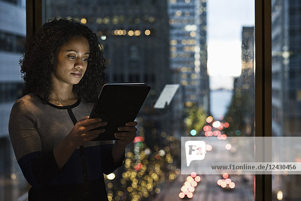 A view of a black businesswoman working on a notebook computer in front of an office window looking out on a city street scene just before dark.