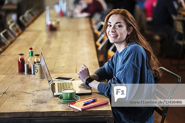 Lächelnde junge Frau mit langen roten Haaren  die am Tisch sitzt und am Laptop-Computer arbeitet.