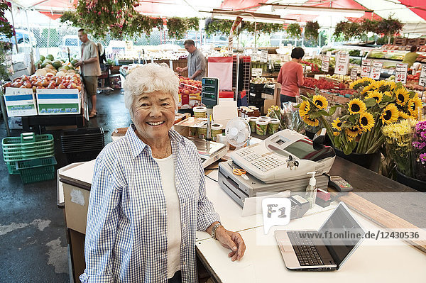 Ältere Frau steht an der Kasse eines Lebensmittel- und Gemüsemarktes und lächelt in die Kamera.