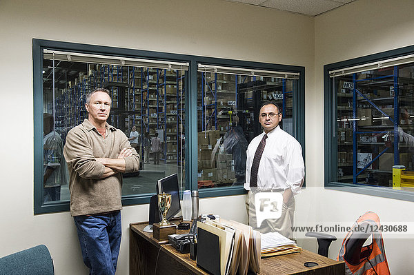Team portrait of a male Hispanic American executive and a male Caucasian factory worker in an office in the middle of a large distribution warehouse full of racks of products stored in cardboard boxes on pallets.