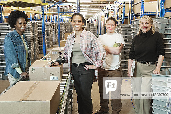 Team portrait of multi-ethnic female warehouse workers working next to a motorized feed conveyor in a large distribution warehouse.