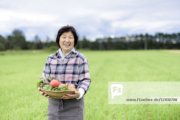 Frau mit schwarzen Haaren in kariertem Hemd steht in einem Feld  hält Korb mit frischem Gemüse und lächelt in die Kamera.